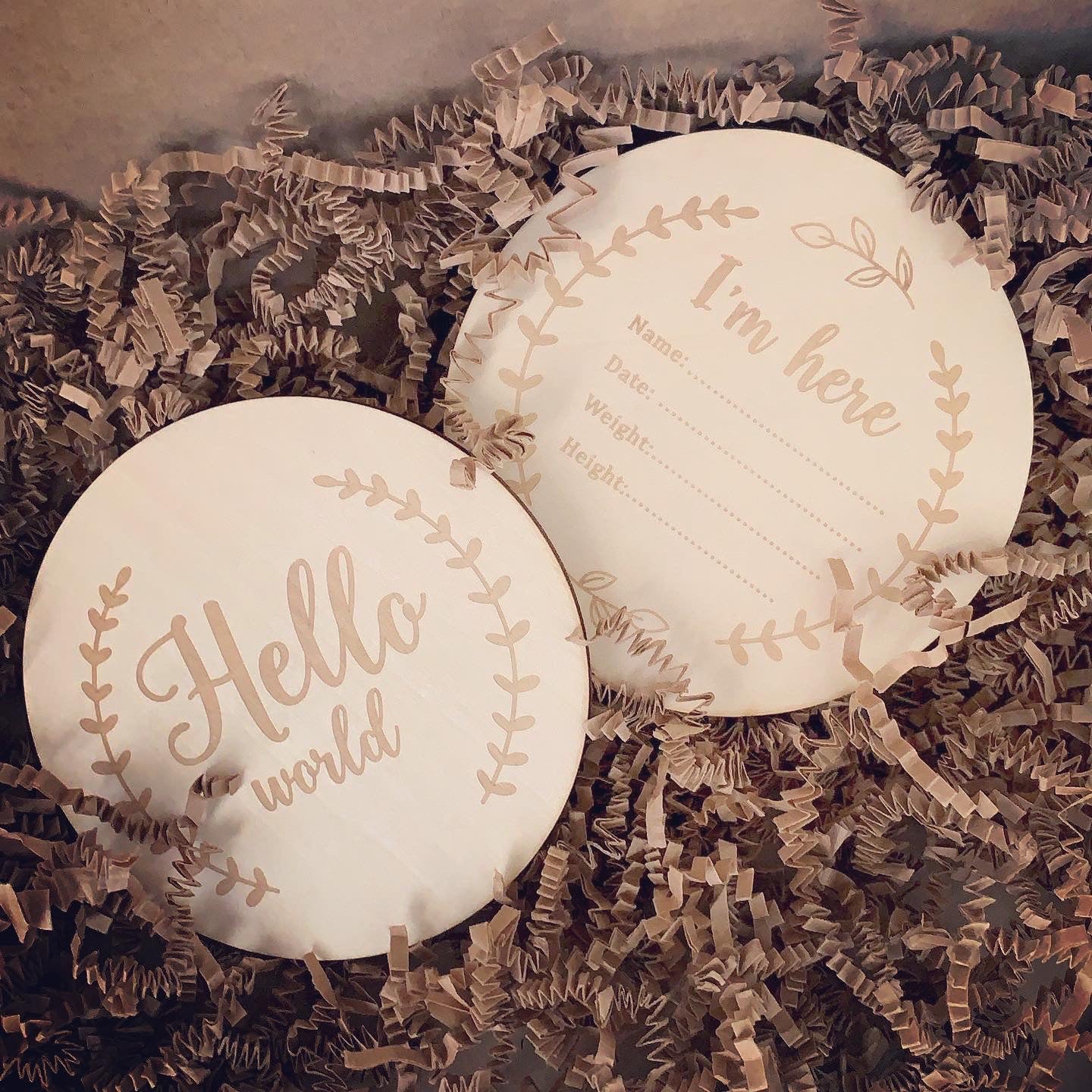 Two sided circled wooden baby shower keepsake, one side decorated with a cut off wreath Hello World in the middle, the second side is also decorated with a wreath around the circle and space to write down birth information